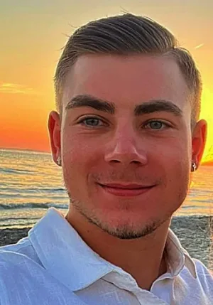 Porträtfoto zur Kundenstimme: junger Mann mit kurzen Haaren und weissem Hemd vor einem Sonnenuntergang am Meer